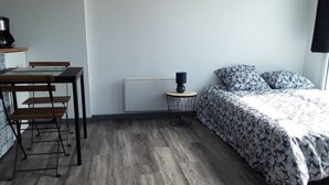 A room with a wooden floor, a bed with patterned bedding, a small round table with a lamp, and a chair. - Studio lumineux proche de la mer (Saint-Nazaire)
