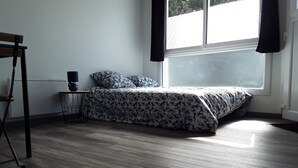 A bedroom with a bed, a small table with a lamp, a chair, and a window with curtains. - Studio lumineux proche de la mer (Saint-Nazaire)