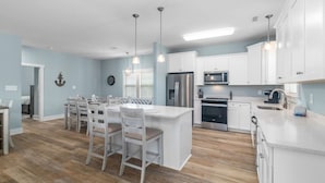A modern kitchen with white cabinetry, stainless steel appliances, and a central island with bar stools. - Modern Beach Villa|2-Min Walk to Beach + Ping Pong (Emerald Isle)