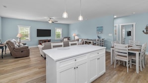 A spacious living area with a large white island, a ceiling fan, and a dining table with chairs. - Modern Beach Villa|2-Min Walk to Beach + Ping Pong (Emerald Isle)
