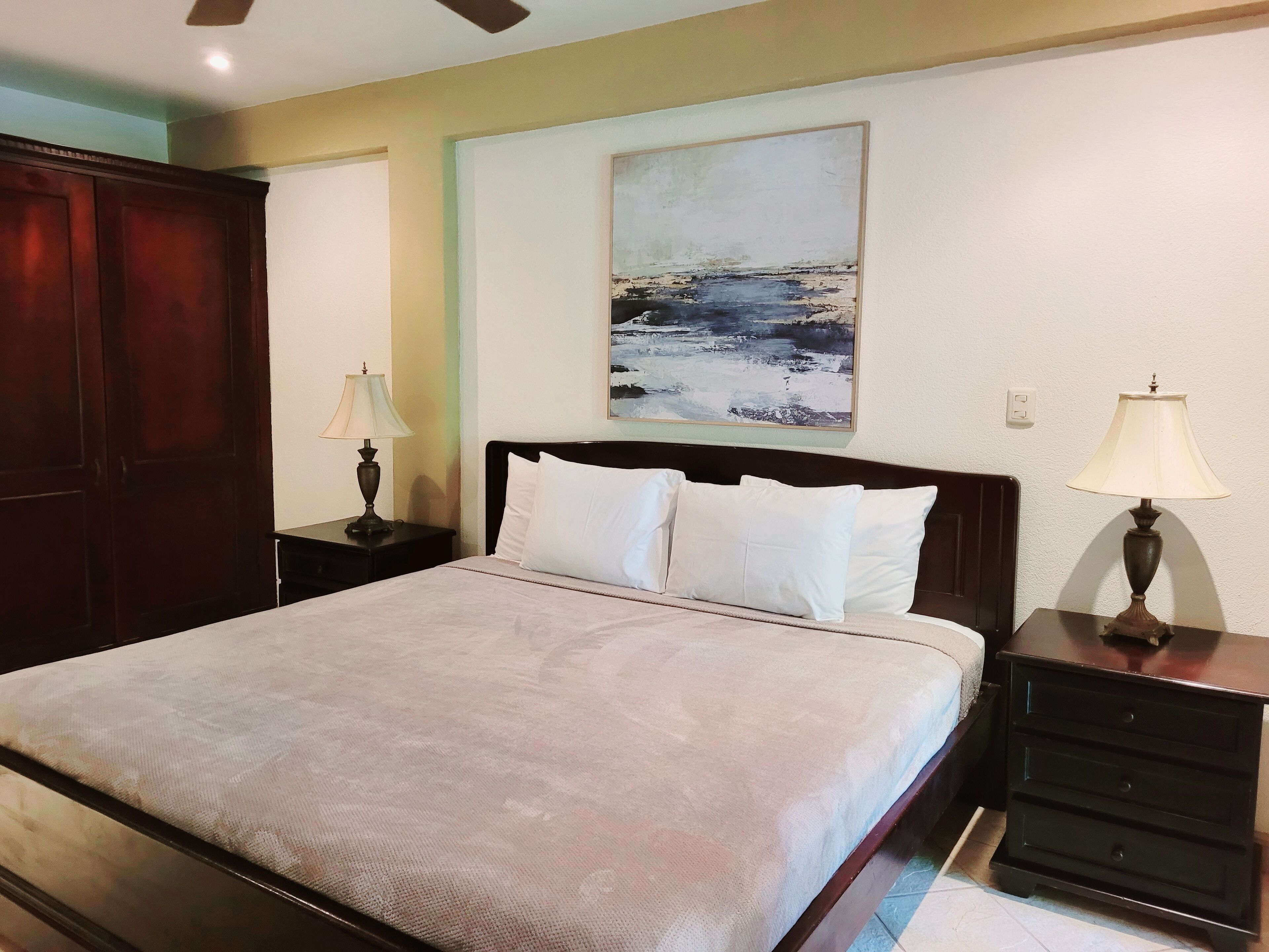 A neatly arranged bedroom with a large bed, two bedside tables with lamps, a wooden wardrobe, and a painting on the wall.