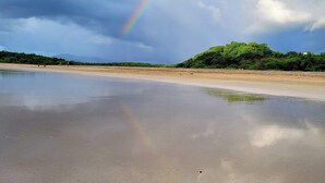 Une plage avec un arc-en-ciel, une eau calme et une colline boisée en arrière-plan.