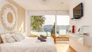 Un dormitorio con cama, televisión montada en la pared, balcón con vistas a palmeras y montañas, y un panel decorativo en la pared.