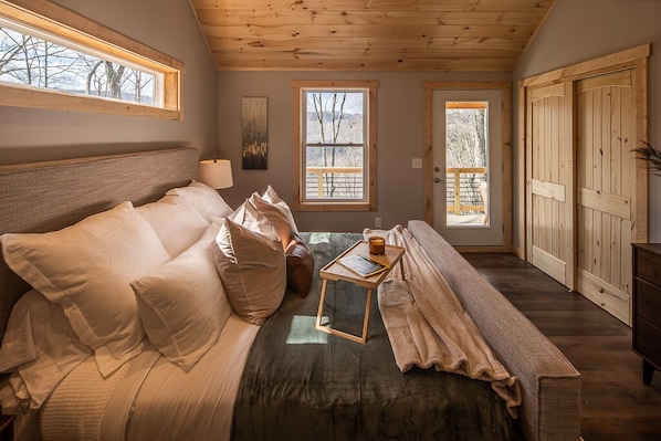 Una habitación acogedora con una cama, una mesita con una vela y un banco. Hay ventanas y una puerta que da al exterior.