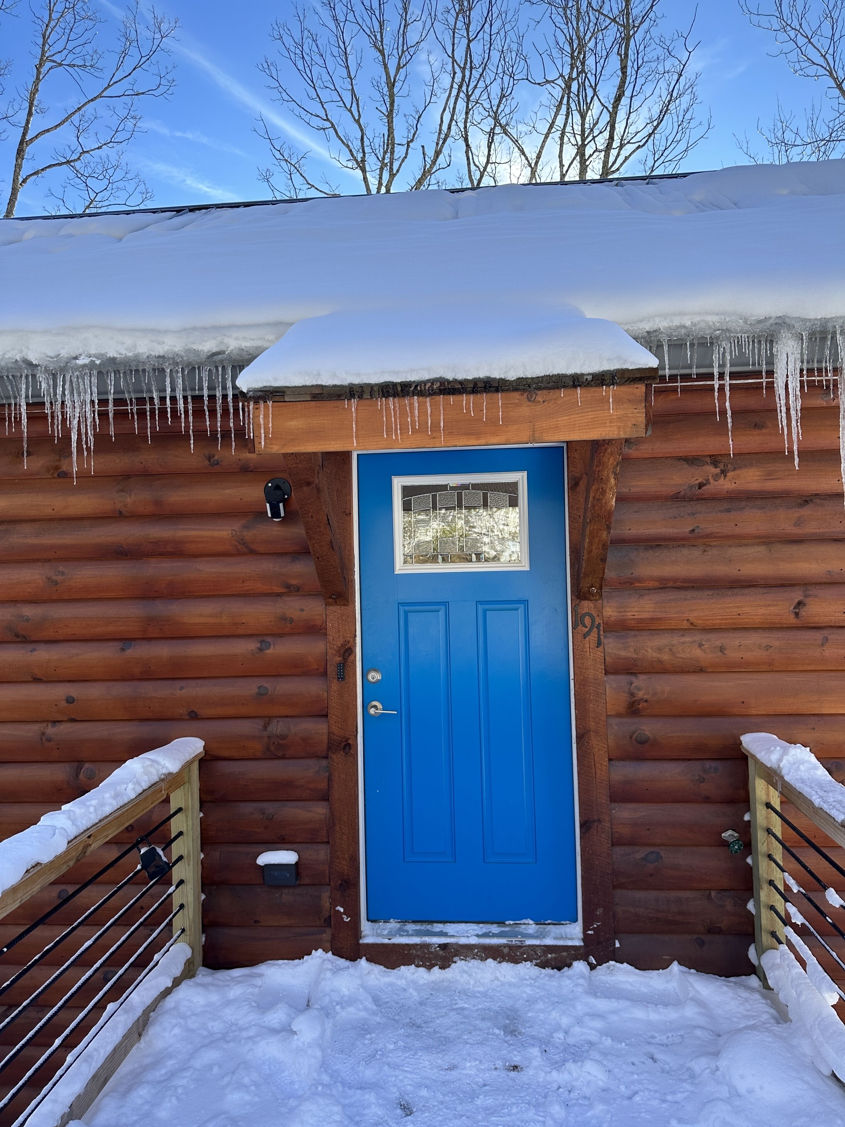 Una cabaña de madera con puerta azul, tejado cubierto de nieve y borbotones de hielo colgando de los aleros.