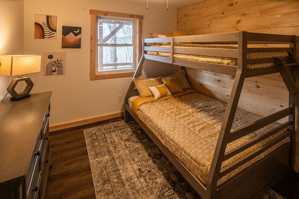 A bunk bed room with a wooden headboard, a desk with a lamp, and a window with a view of trees.
