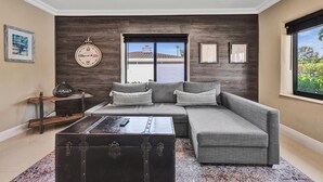 A modern living room with a grey sectional sofa, a dark wooden accent wall, a large window with a view, and a vintage suitcase on a rug. - Romantic Retreat w/ Private Pool. Near Las Olas (Fort Lauderdale)