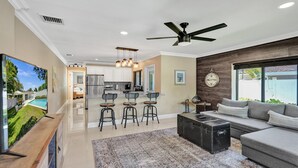 A modern living room with a grey sofa, a coffee table, and a ceiling fan. There is a kitchen area with white cabinets and a dining space with bar stools. - Romantic Retreat w/ Private Pool. Near Las Olas (Fort Lauderdale)