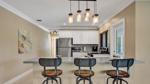 A modern kitchen with white cabinets, a stainless steel refrigerator, a microwave, and a coffee maker. There are three bar stools with wooden seats and metal frames. - Romantic Retreat w/ Private Pool. Near Las Olas (Fort Lauderdale)