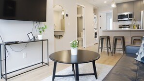 A modern living room with a flat-screen TV, a round table with a vase, a sofa, and a kitchen area with a microwave and stools. - Enchanted Mountain Getaway: Accessible Retreat in Canmore, Close to Banff (Canmore)