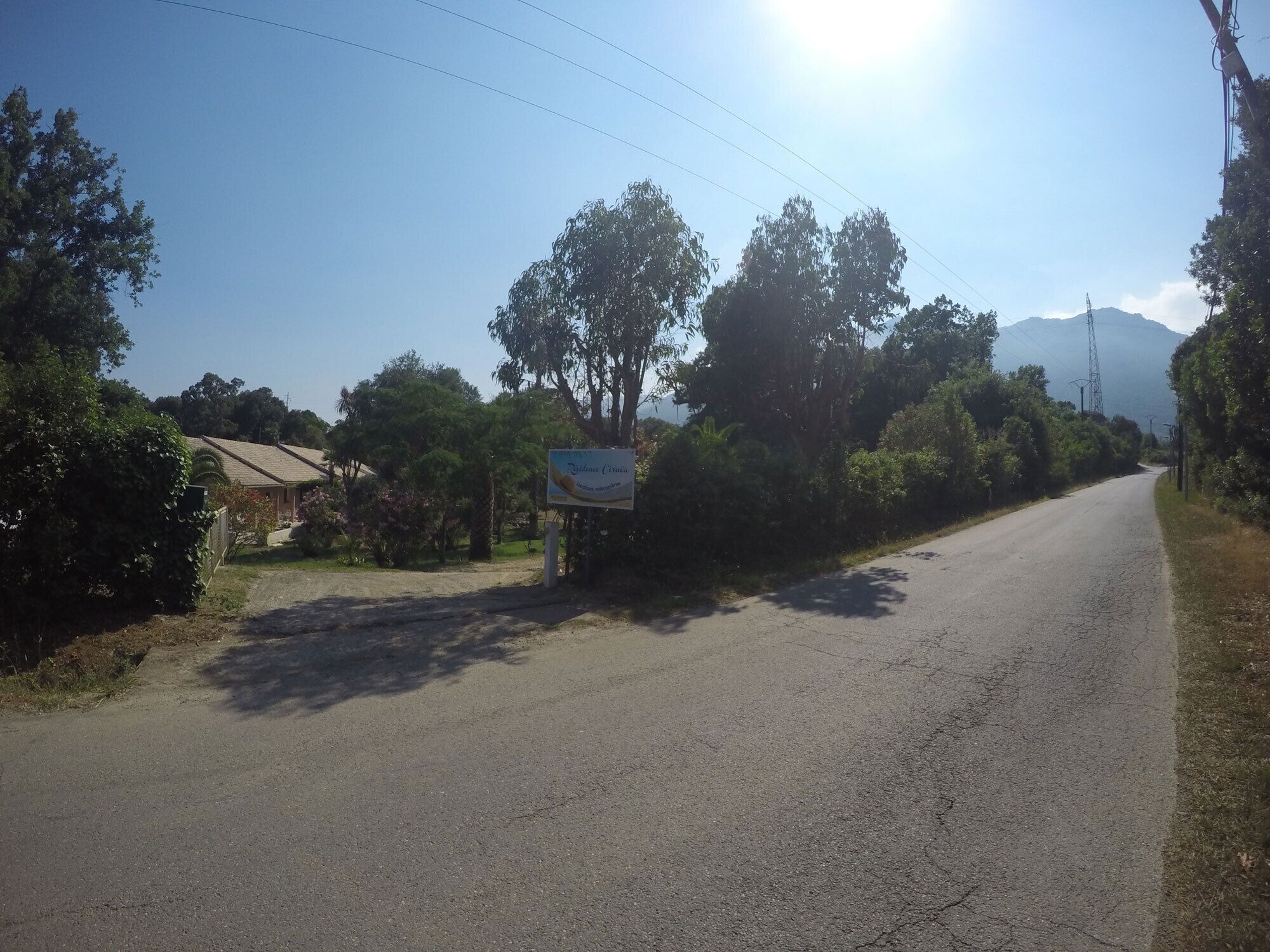 A rural road with a signboard, surrounded by greenery and a distant mountain.