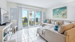 A spacious living room with a large sofa, a flat-screen TV, and a balcony with sliding glass doors. - Ocean View Condo Close to the Pier on the Beach! (Cocoa Beach)