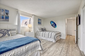 Une chambre avec deux lits, une table de chevet, une lampe, un tableau décoratif au mur et une porte donnant sur une autre pièce.