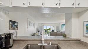 A modern kitchen with a double sink, a coffee maker, and a view to a living area with a sofa and a balcony. - Ocean View Condo Close to the Pier on the Beach! (Cocoa Beach)