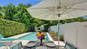 A poolside dining area with a table set for four, surrounded by a white fence, and a large umbrella providing shade. - Tranquil Getaway w/ Private Heated Pool (Fort Lauderdale)