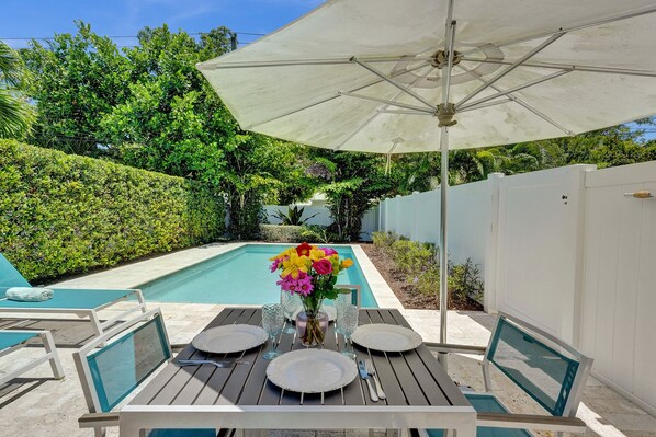 A poolside dining area with a table set for four, surrounded by a white fence, and a large umbrella providing shade.