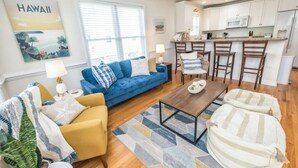 Ein modernes Wohnzimmer mit einem blauen Sofa, einem gelben Sessel und einem gestreiften Hocker. Ein Holztisch und ein gemusterter Teppich befinden sich in der Mitte. Im Hintergrund ist eine Küche mit weißen Schränken und einer Bar zu sehen.
