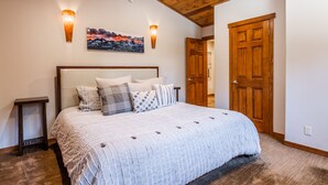 A bedroom with a large bed, wooden ceiling, two wall-mounted lights, a painting, and a wooden door.