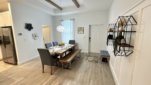 Un espace repas moderne avec une table en bois, des chaises et un banc. On aperçoit une cuisine sur la gauche, une porte au fond et un élément décoratif mural sur la droite.