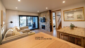 A modern lounge area with a view of the lake, a stone fireplace, and a dining table. -  Lake Front Living New and Cosy seperate ground floor appartment (East Jindabyne)