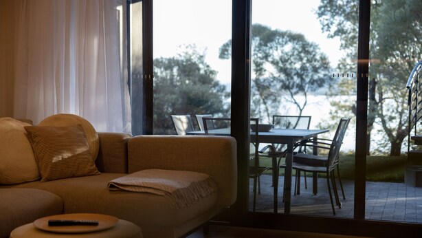 A modern living room with a sofa, ottoman, and a dining area visible through large glass doors. -  Lake Front Living New and Cosy seperate ground floor appartment (East Jindabyne)