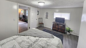 Quarto com cama, cômoda, cadeira, televisão e janela.