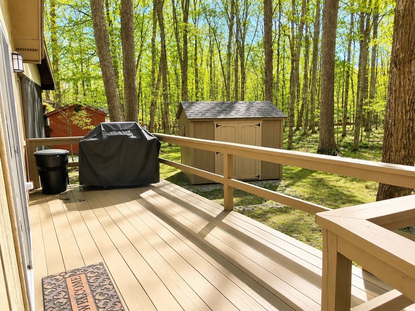 A wooden deck with a railing, a barbecue grill, a trash can, and a shed in a wooded area.