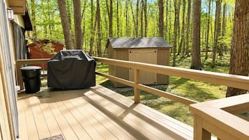 A wooden deck with a railing, a barbecue grill, a trash can, and a shed in a wooded area.