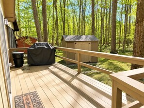 A wooden deck with a railing, a barbecue grill, a trash can, and a shed in a wooded area. - Beautiful Lodge in The Hideout!! (Lake Ariel)
