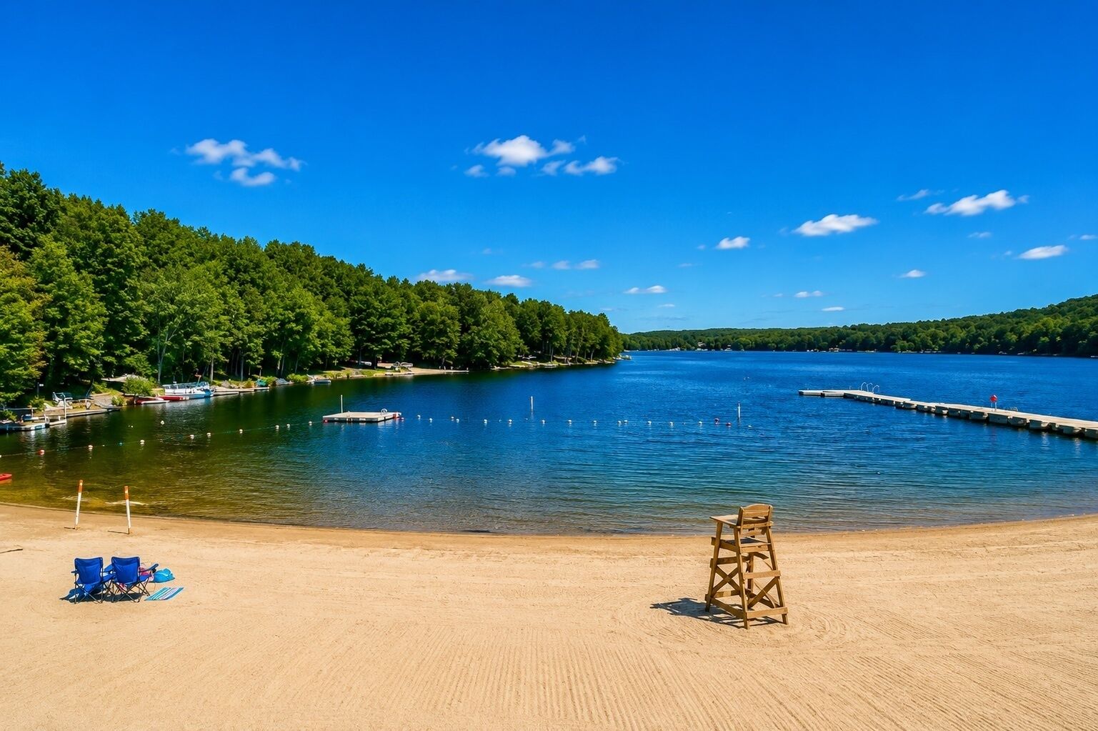 A sandy beach with a lifeguard chair, a clear blue sky, and a calm lake surrounded by trees.