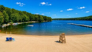 A sandy beach with a lifeguard chair, a clear blue sky, and a calm lake surrounded by trees. - Beautiful Lodge in The Hideout!! (Lake Ariel)
