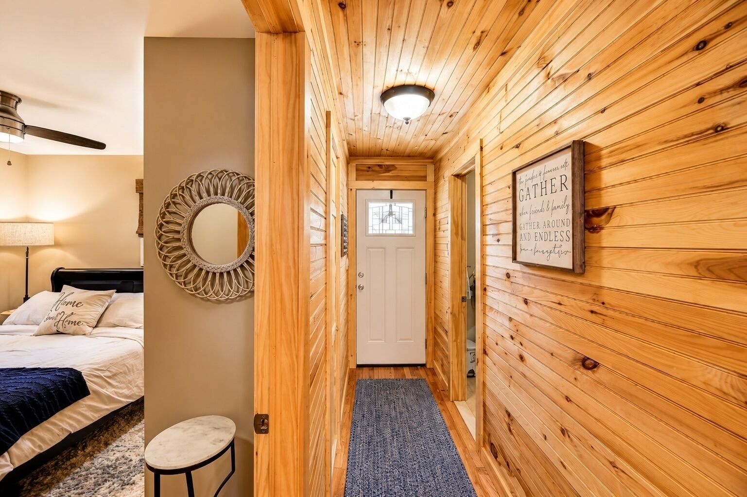 A wooden interior hallway with a bed, a round mirror, a framed sign, and a door.