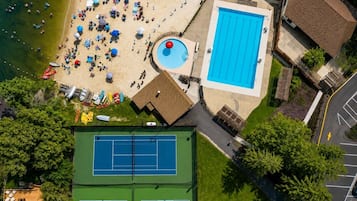 An aerial view of a beach area with a swimming pool, a tennis court, and a building with a roof.