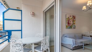 Un balcó amb taula i cadires blanques, un sofà i una porta de vidre que dona a una habitació interior amb un quadre.