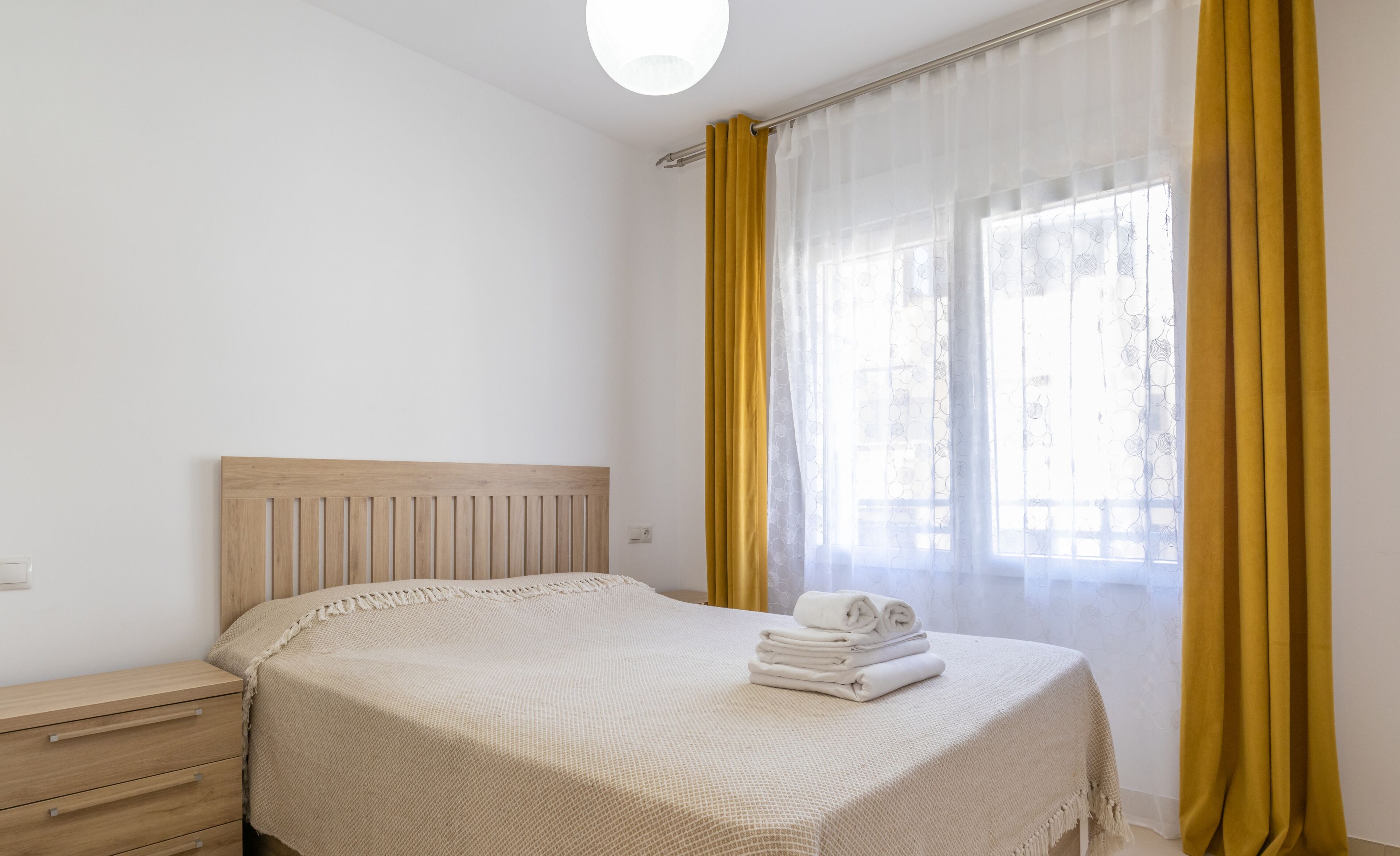 Une chambre à coucher avec une tête de lit en bois, un lit recouvert d’une couverture beige à texture, une table de chevet avec une lampe et une fenêtre ornée de rideaux jaunes.