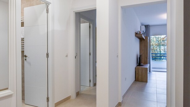 Passadís amb parets blanques i portes que donen a diferents habitacions. Hi ha una banca de fusta a la cantonada i es veu l'exterior a través d'una finestra.