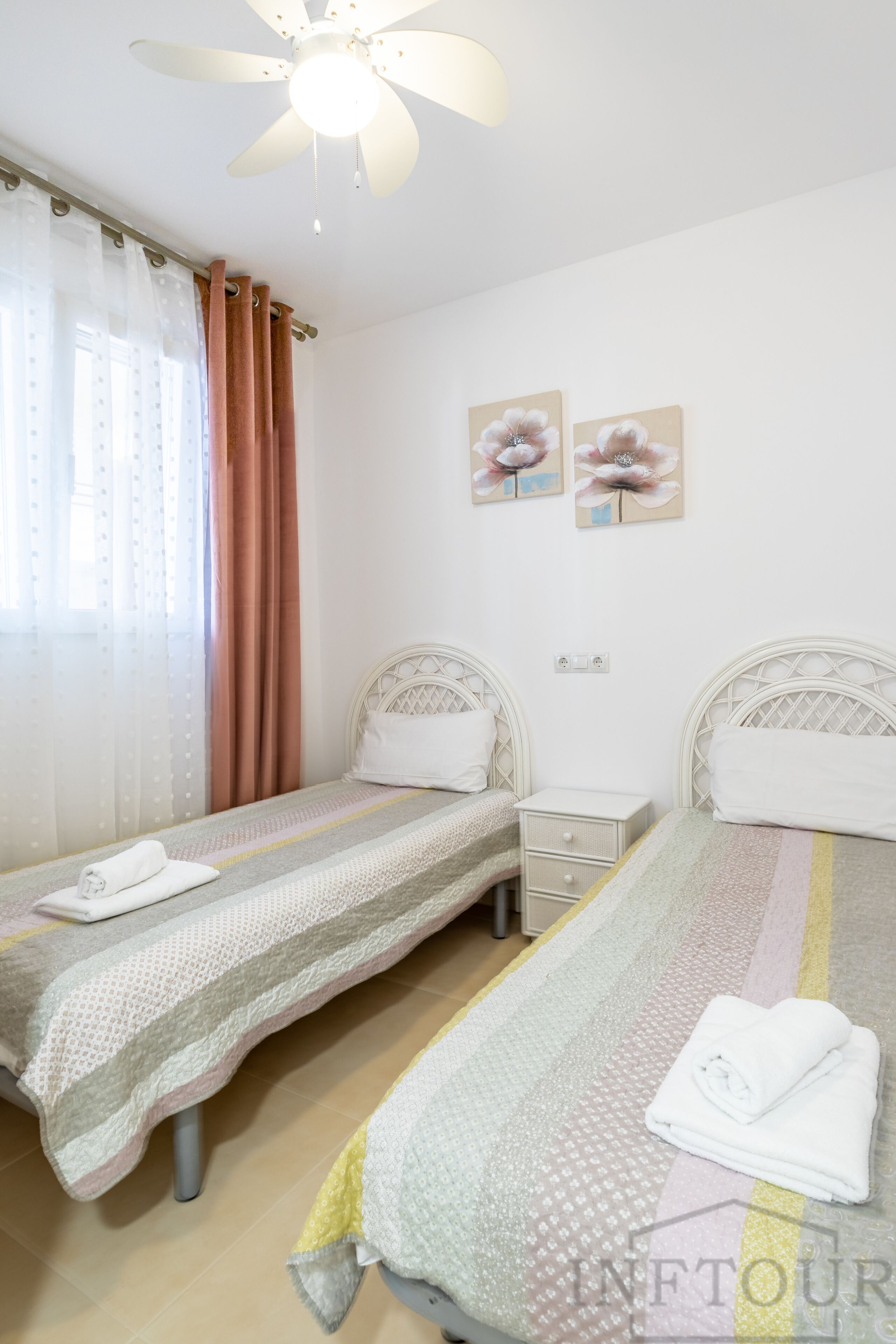 Une chambre avec deux lits simples, un ventilateur de plafond, une table de chevet et une fenêtre avec des rideaux.