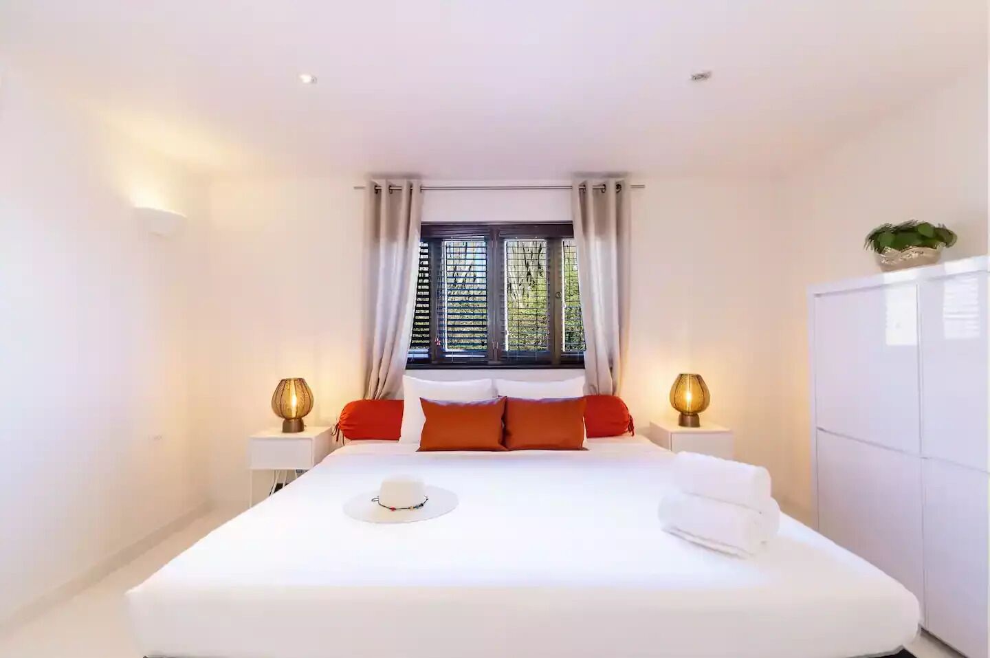 A neatly arranged bedroom with a large bed, two bedside lamps, a window with curtains, and a small plant on the dresser.