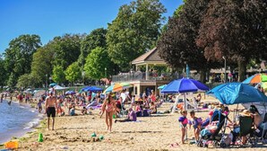A crowded beach with people sunbathing and walking, umbrellas, and a pavilion in the background. - Luxury Property-Walk to lake and downtown Lake Geneva. Pet Friendly, Sleeps 8 (Lake Geneva)