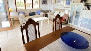 Ein Wohnzimmer mit einer Couch, einem Esstisch und einem Sessel. Es gibt einen Deckenventilator, ein Fenster mit Jalousien und ein Bücherregal.