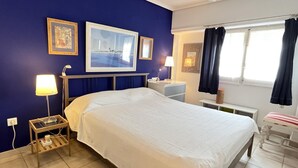 Ein Hotelzimmer mit einem großen Bett, einem Nachttisch mit Lampe, einem kleinen Schreibtisch mit Stuhl, einem Fenster mit Vorhängen und Bildern an den Wänden.
