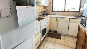Eine kompakte Küche mit weißen Schränken, einem Kühlschrank, einem Geschirrspüler und einer Mikrowelle. An der Wand hängt ein Kalender, und ein Fenster lässt Tageslicht herein.