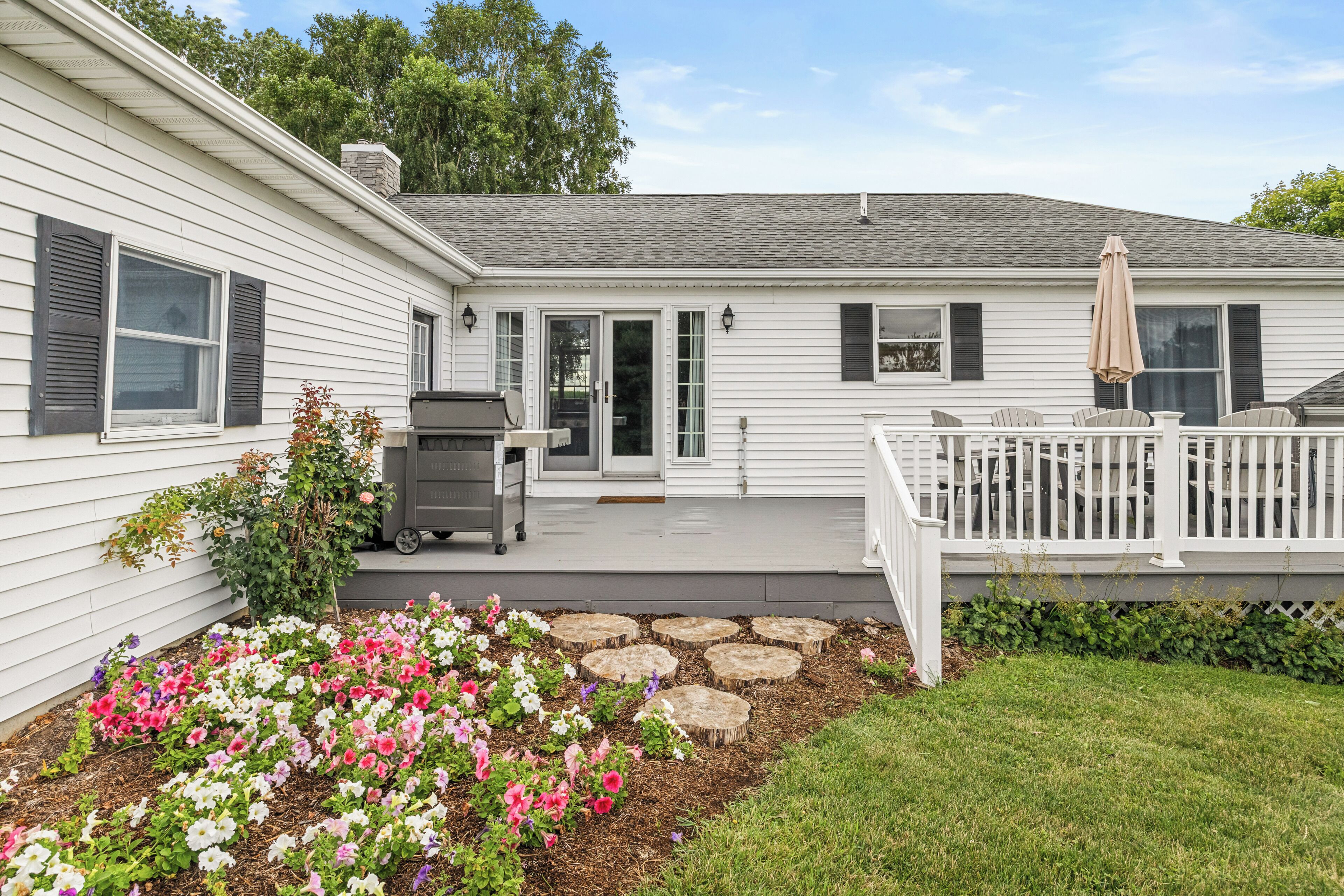 A white house with a covered patio, a grill, and a flower bed.