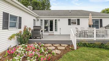 A white house with a covered patio, a grill, and a flower bed.