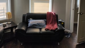 A living room with a black leather sofa, a red throw blanket, and a blue pillow. There is a window with blinds, a lamp on a side table, and a door to the right. - Irvington Apartment (Irvington)