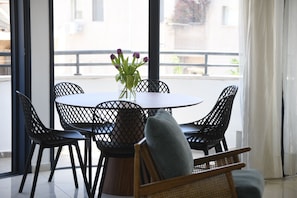 A dining area with a round table and six chairs, a vase with flowers, and a view of the outside through large windows.