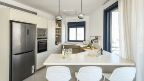 Eine moderne Küche mit einem Edelstahl-Kühlschrank, einer Mikrowelle und einem eingebauten Backofen. Drei weiße Stühle stehen um eine zentrale Kochinsel mit Spüle und einer Weinflasche.