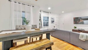 A modern living room with a wooden dining table, bench, grey sofa, and a large window with sheer curtains.