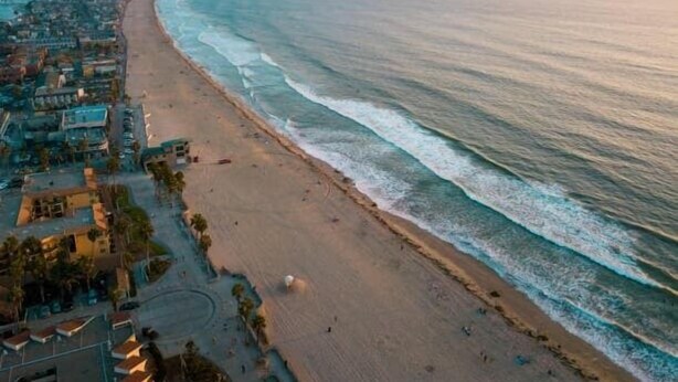 An aerial view of a beach with a long stretch of sand, a few buildings, and a clear sky.