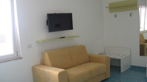 A room with a beige sofa, a flat-screen TV mounted on the wall, a small white table, and a window with a view. - Studio with equipped kitchen - Pisciotta (Pisciotta)